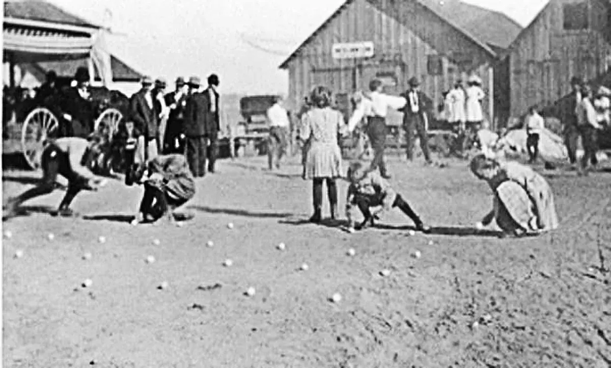 Children crouched over a game of marbles in a dirt street, with a crowd, a wagon and wooden buildings behind them