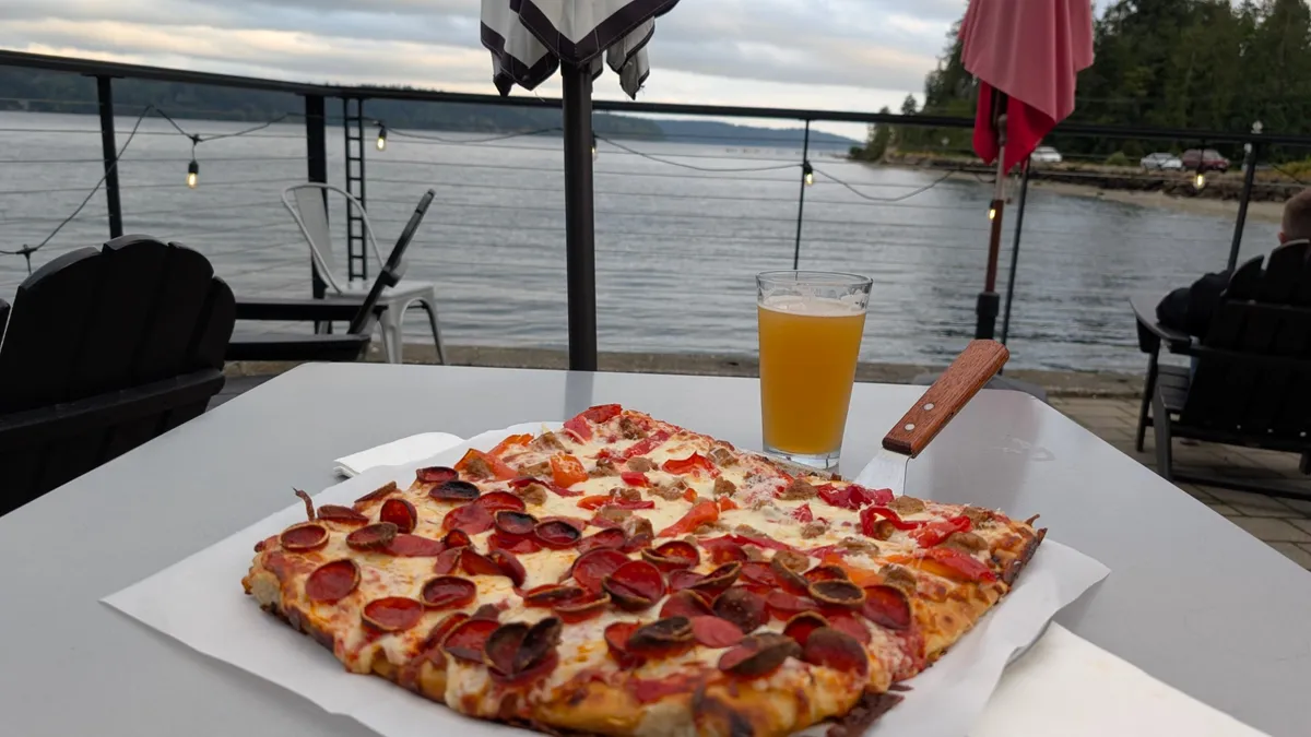 A pepperoni sourdough pizza and a hazy pint on a patio table, the bay and string lights behind
