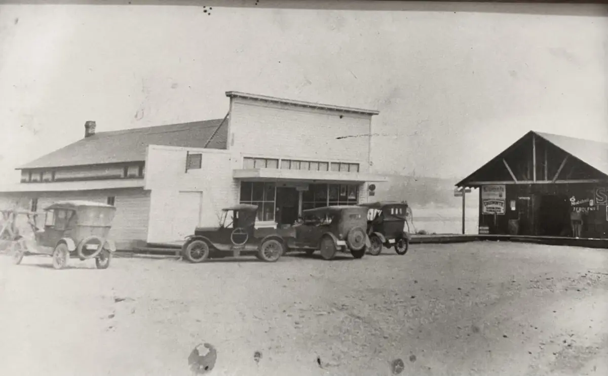 Black and white photo of the store with early automobiles parked out front and a Mobiloil sign on the shed next door